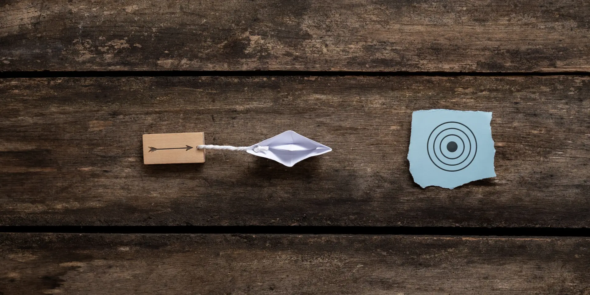 Origami boat pulling wooden peg with arrow drawn on it towards a blue paper with target sign. Conceptual image of vision and clear goals. Over rustic wooden background.