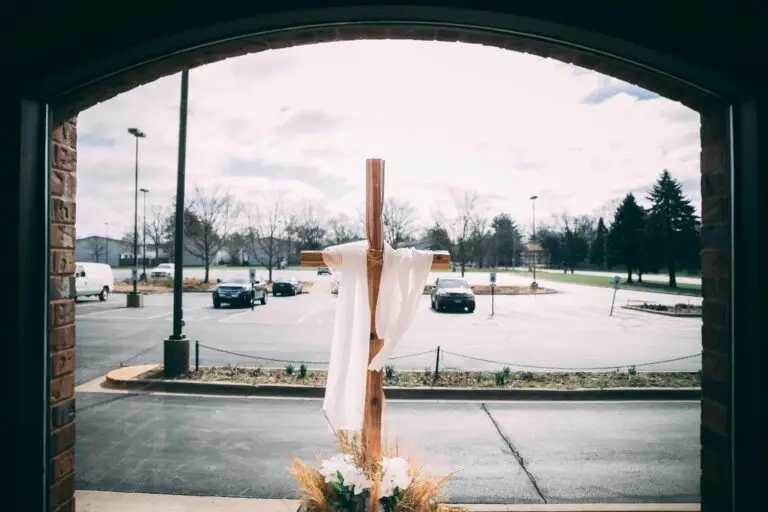 Wooden cross with white strip of cloth wrapped around it on sidewalk with parking lot in background