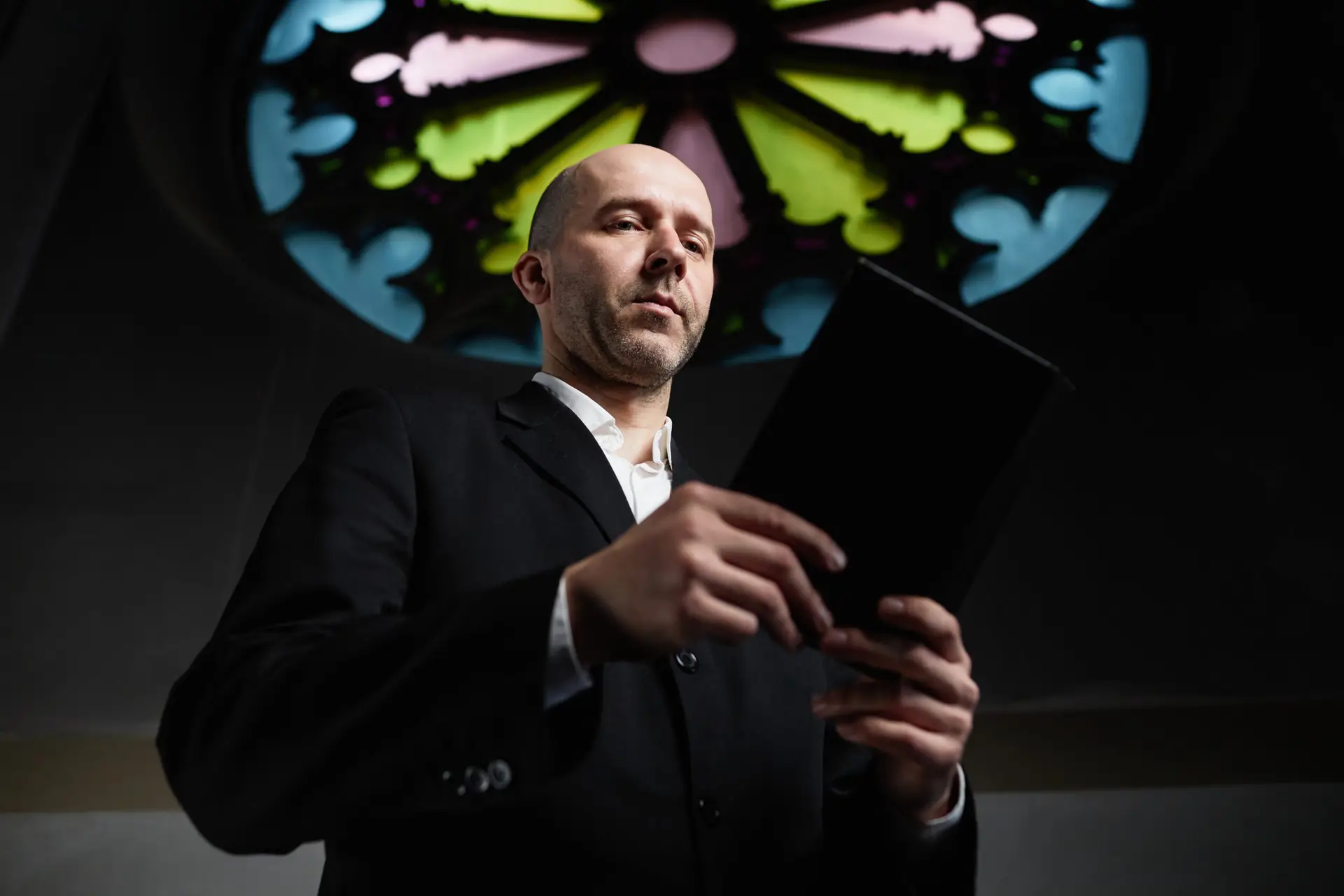 Low angle view of man in black suit holding Bible standing in old church with ornate of stained glass in background