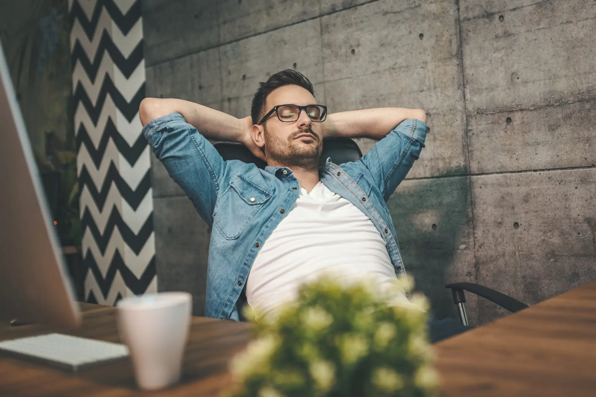 Young man is resting leaning back in office chair with eyes closed and arms behind head