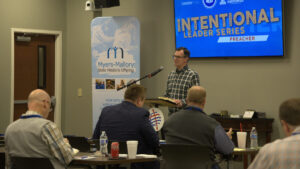 Herb Reavis stands in front of a small podium in front of a group of men, with Intentional Leader Series: Preacher graphic on a TV screen behind him.