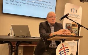 Tim Beougher stands behind a podium in front of a TV screen and speaks to participants at an Intentional Leader event